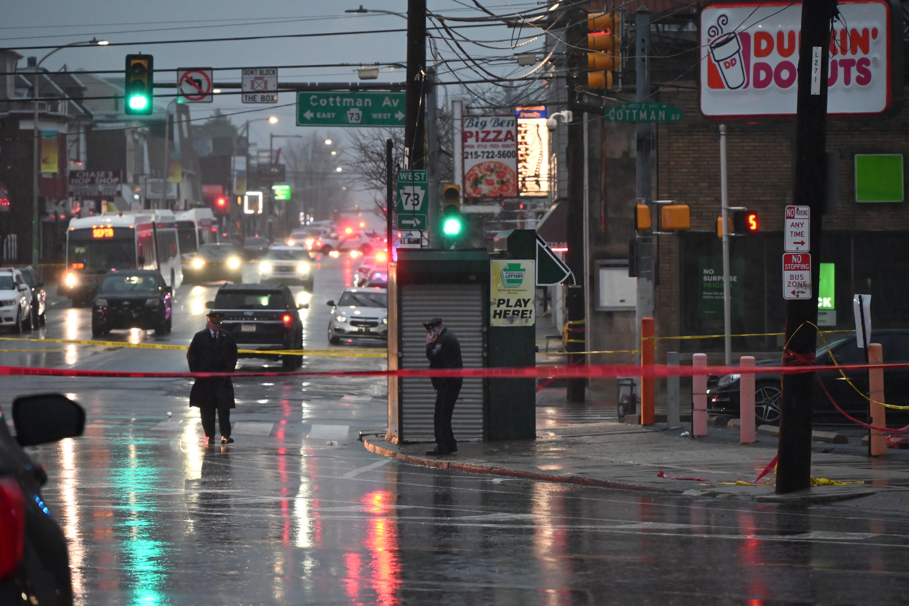 Two police officers stand on the sidewalk and in the street with a red caution tape across the street with cars in the background.