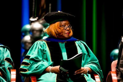 A Black woman wearing Doctorate graduation regalia speaks sits on stage during a graduation ceremony.