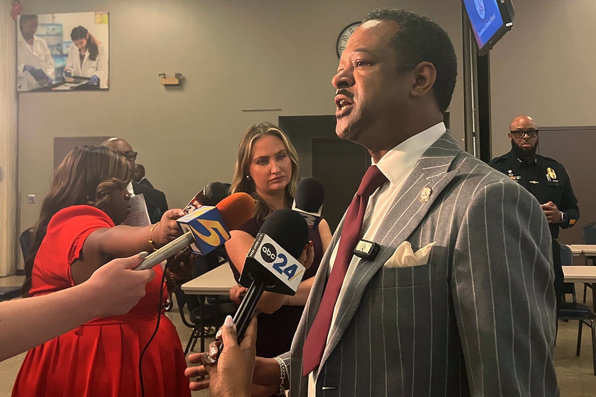 A man in a suit jacket speaks to people holding microphones.