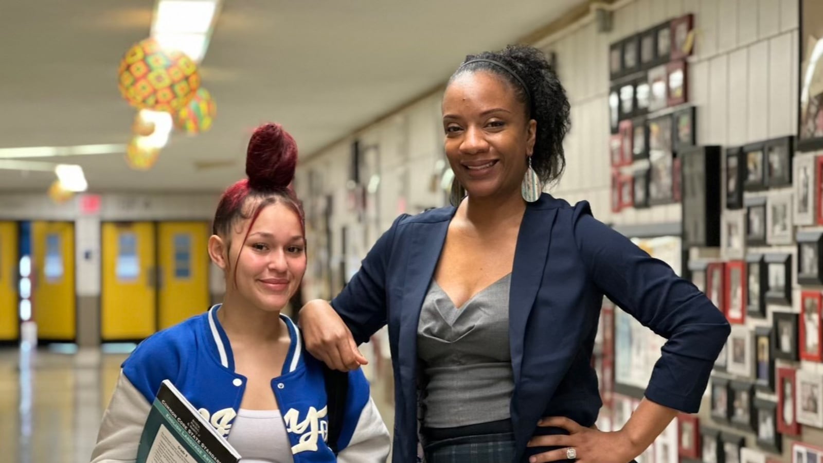 A student and a teacher stand together in a school hallway and pose for a photo.