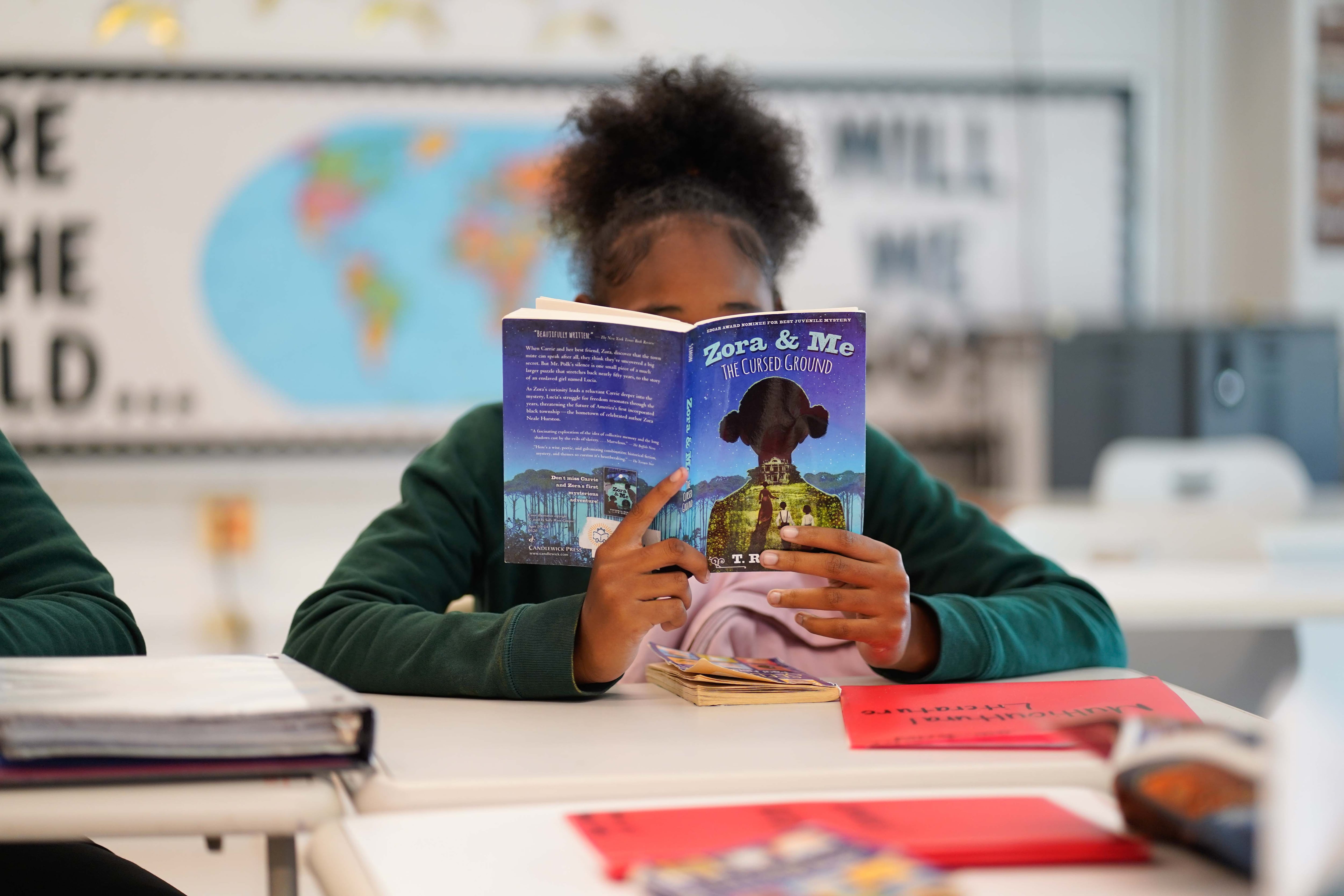 A student with dark hair holds a book covering their face while sitting at their desk. The book reads "Zora and Me the Cursed Ground."