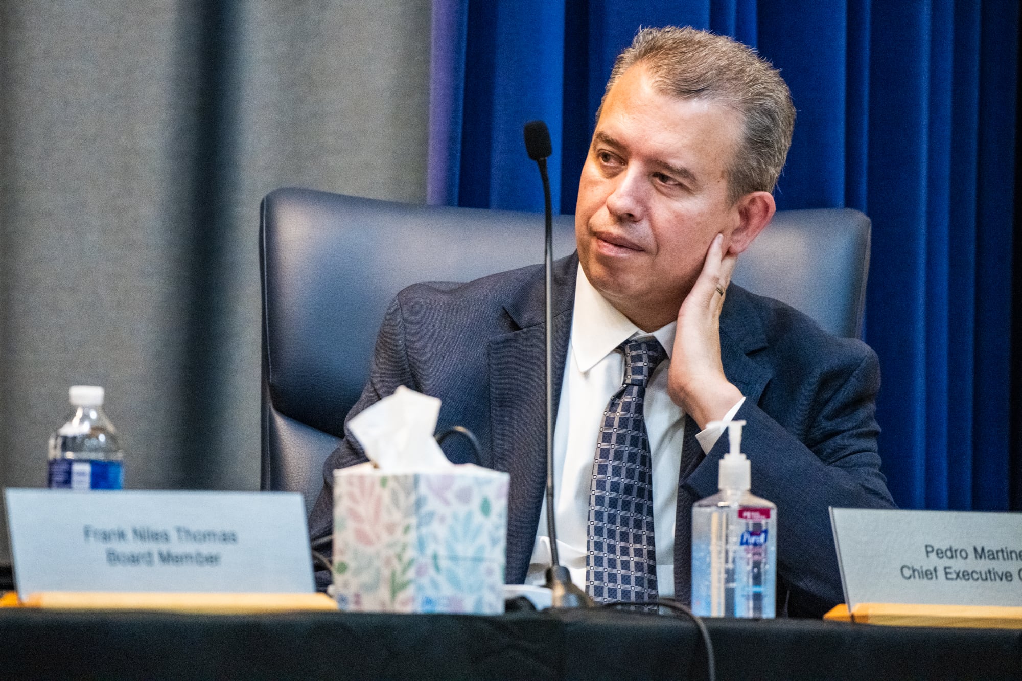 A man speaks from a microphone on a wooden desk.