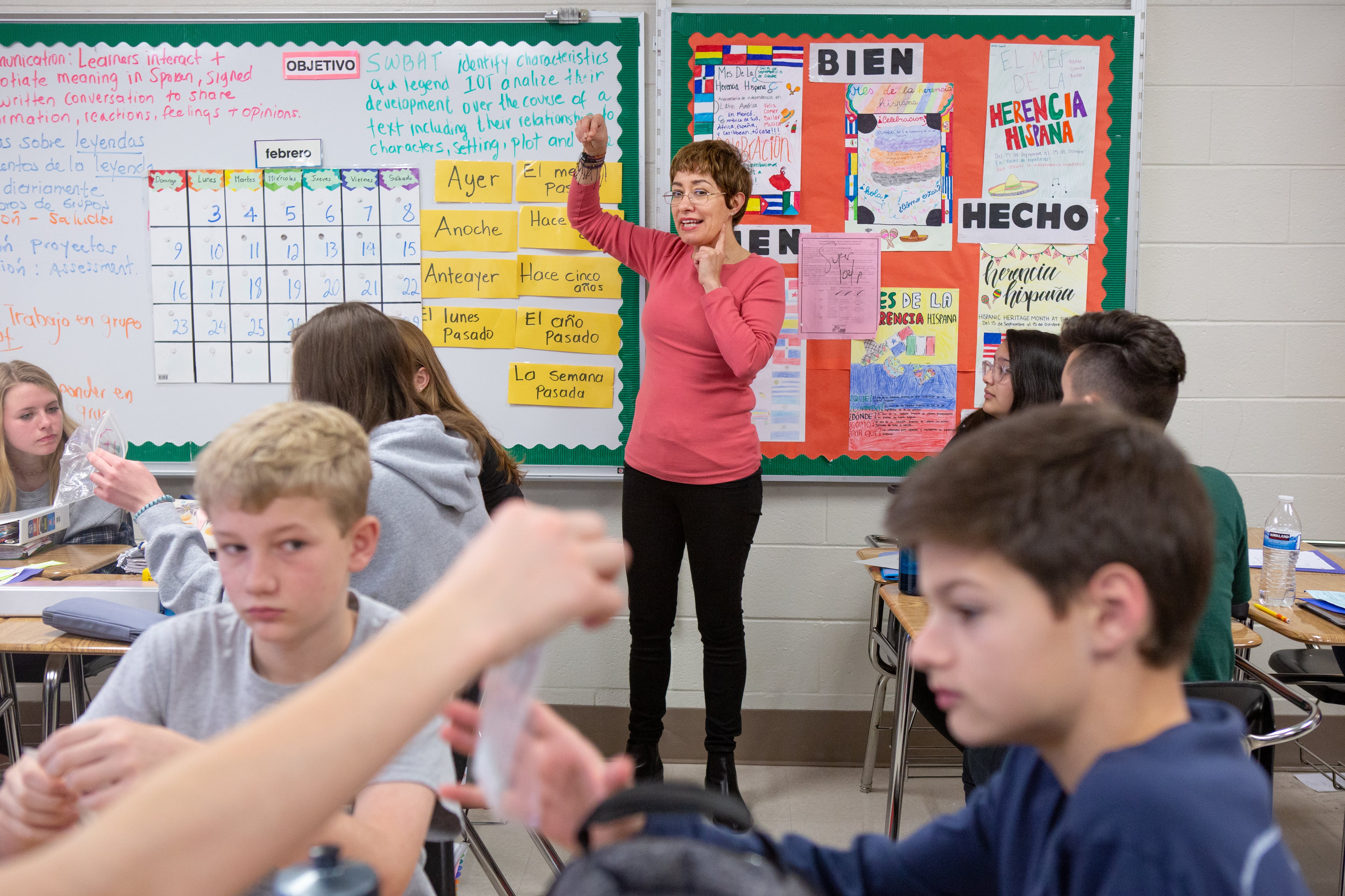 Students sit at desks in a classroom while a teacher provides instruction. A bulletin board behind her is covered with Spanish vocabulary words such as ayer, anoche, bien, and lunes.