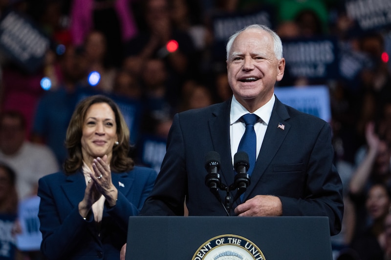 A man in a dark suit speaks from a podium with the Vice President and a large crowd of people.