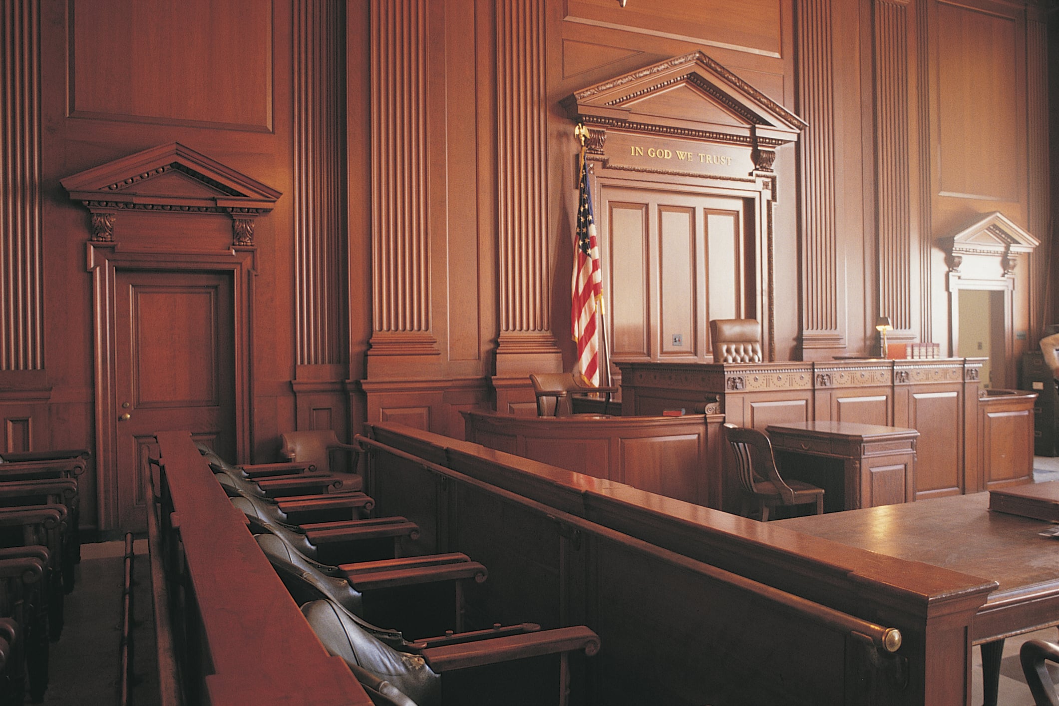A courtroom sits empty, with deep brown walls and an American flag standing behind the judge’s bench.