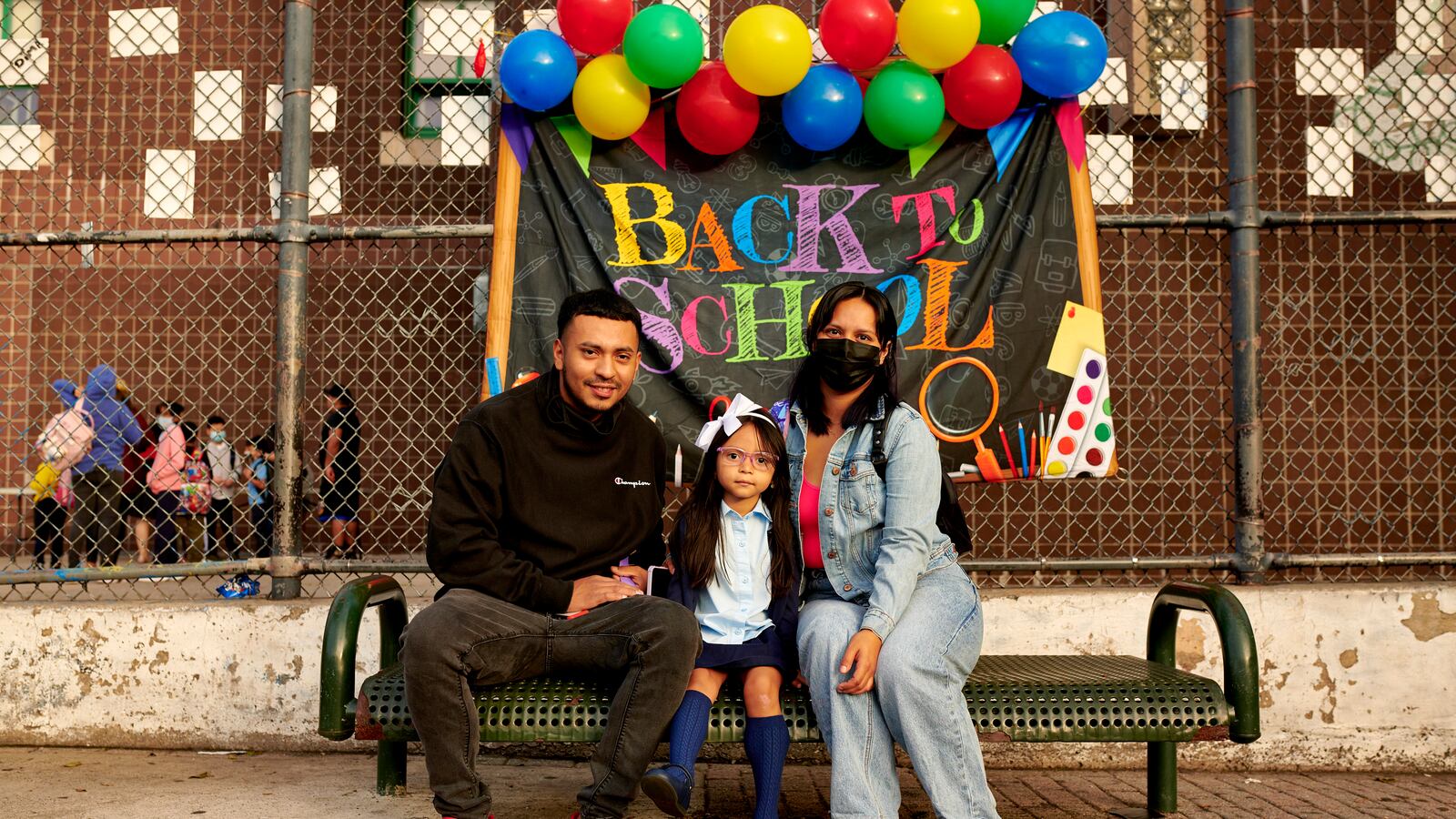 Two parents pose with their daughter for a portrait in front of a banner that reads, “Back to School.”