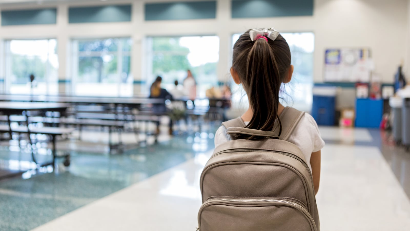 A young schoolgirl with dark hair and a tan backpack pauses as she walks into a school cafeteria.