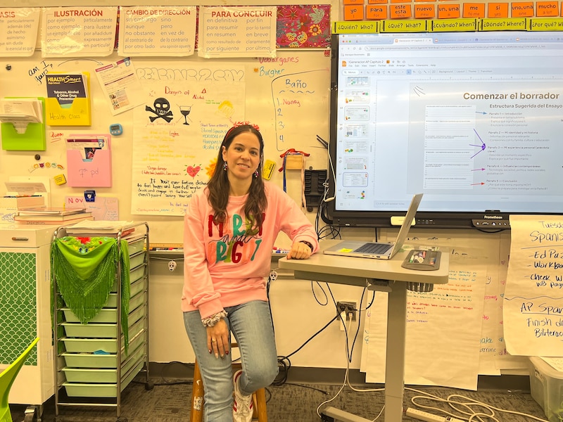 A woman with brown hair who is wearing a pink tshirt and jeans sits to the left of a desk. To her right is a display board and behind her are a lot of things on a bulletin board