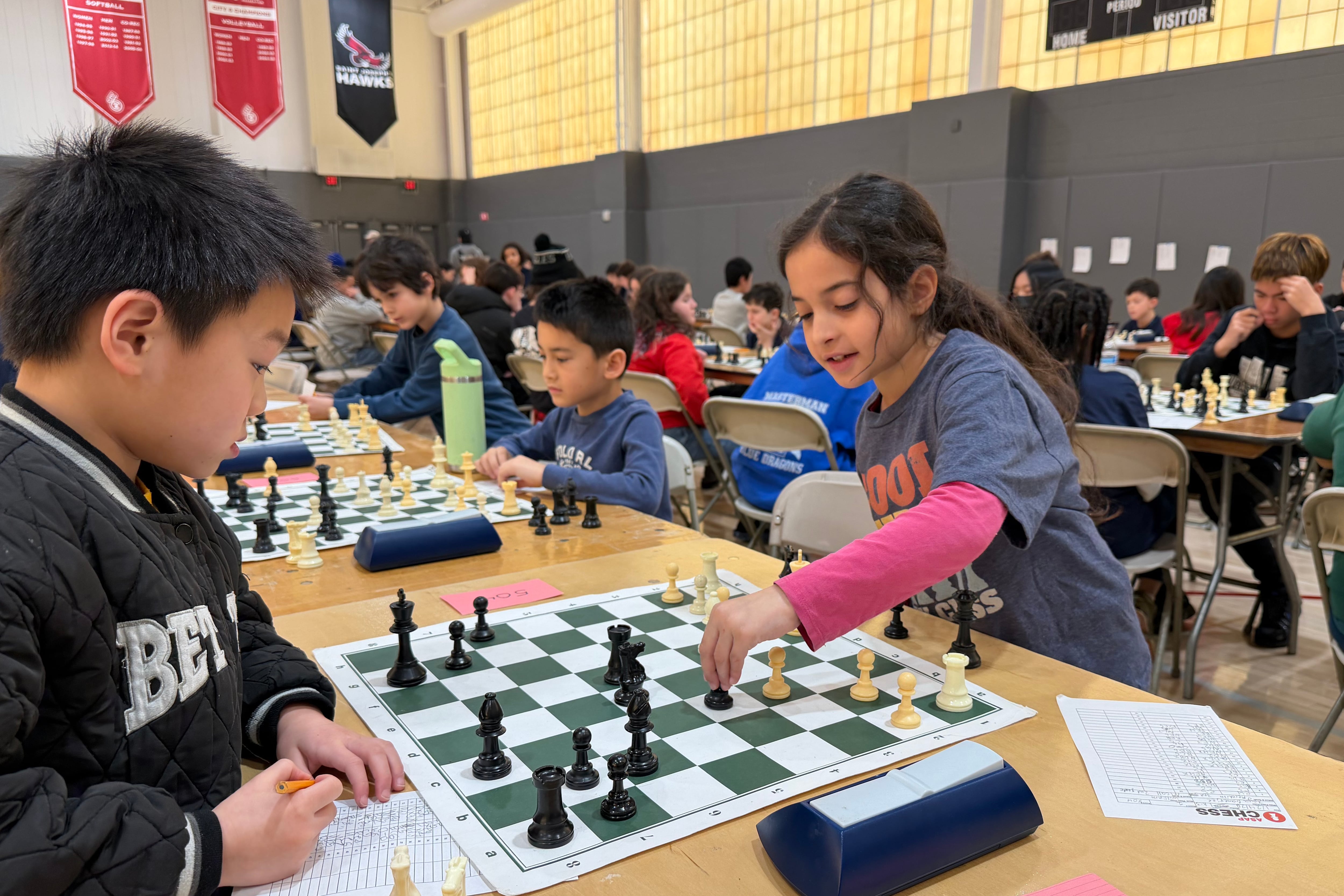 A photograph of two young students playing chess in a large room with other students playing chess in the background.