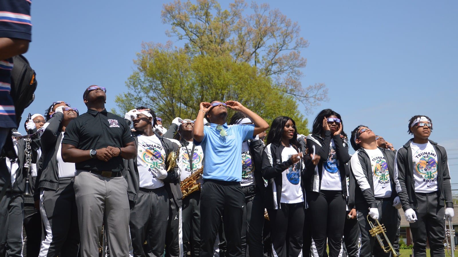 A group of high school students all wearing eclipse viewing glasses stand in a line outside looking up at the sky.