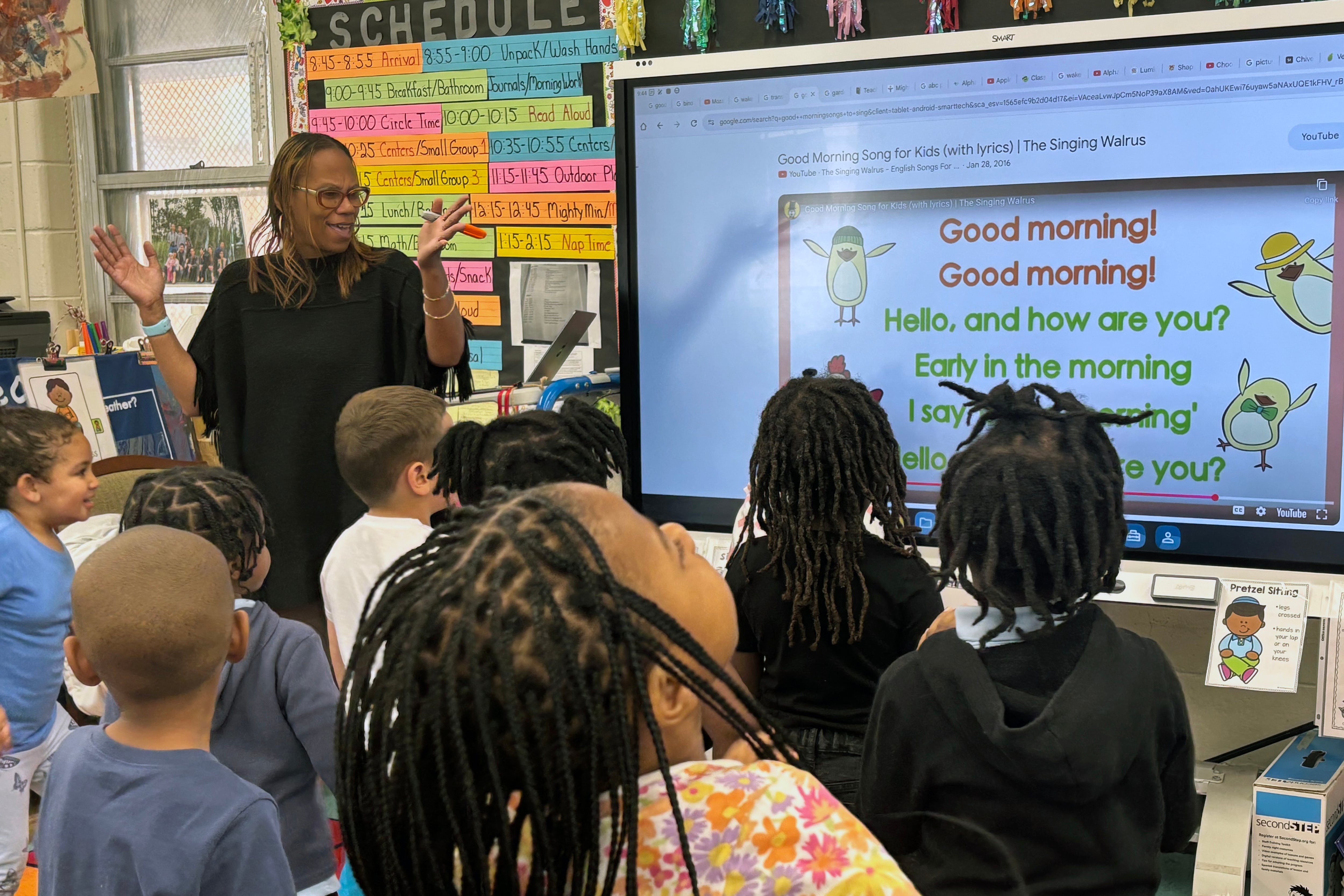 A Black woman teaches a class full of young students in a colorful classroom.