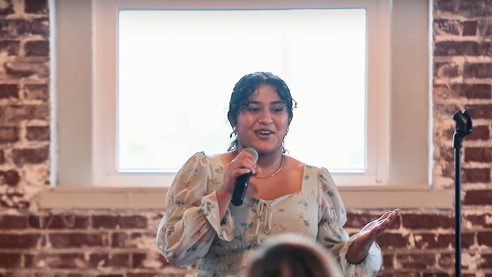 Shreya Ganesh stands before a crowd in a white shirt and is holding a microphone. She stands before a brick wall.