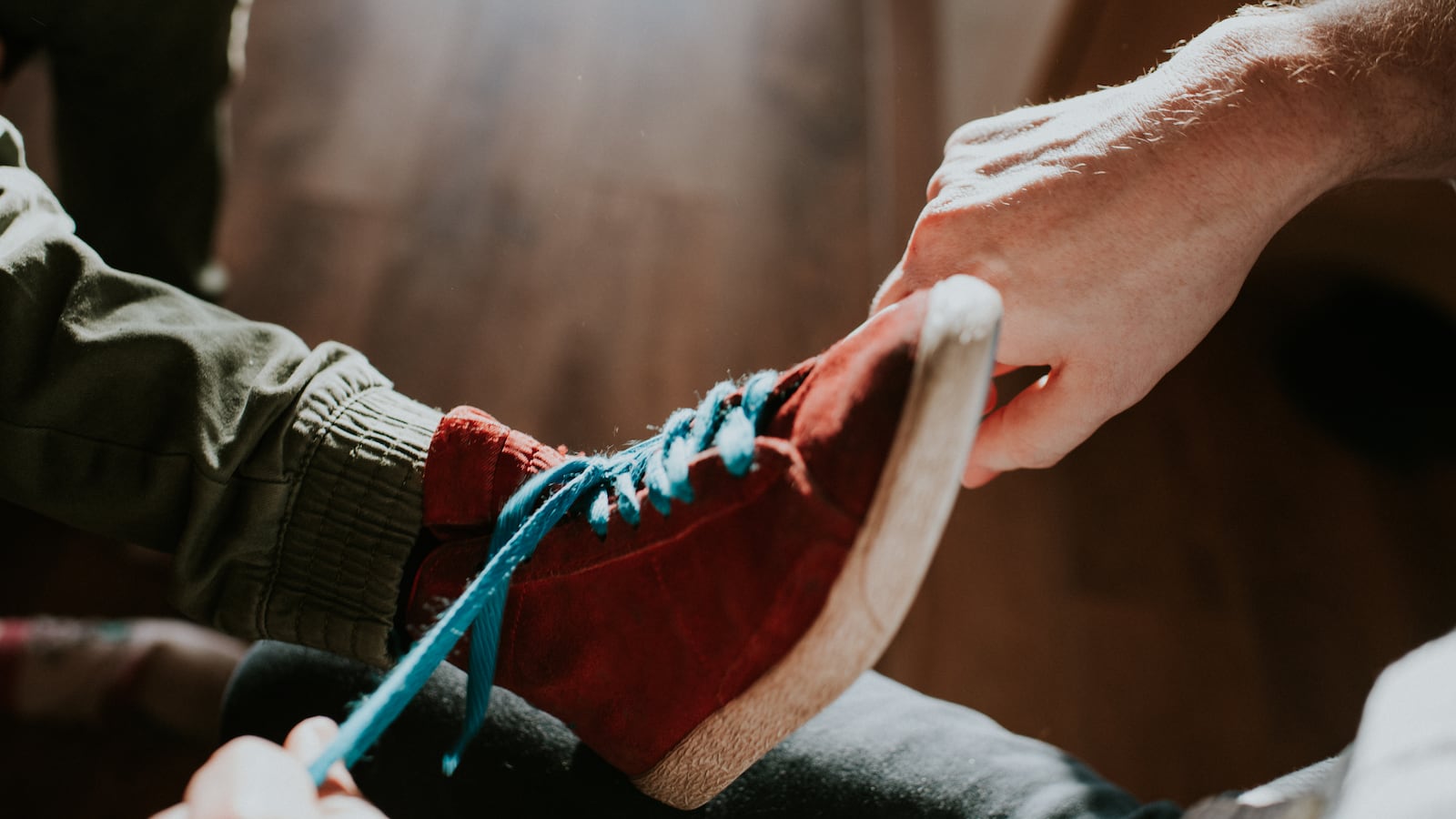 An adult tying a kid’s shoe. The shoe is red with blue laces.