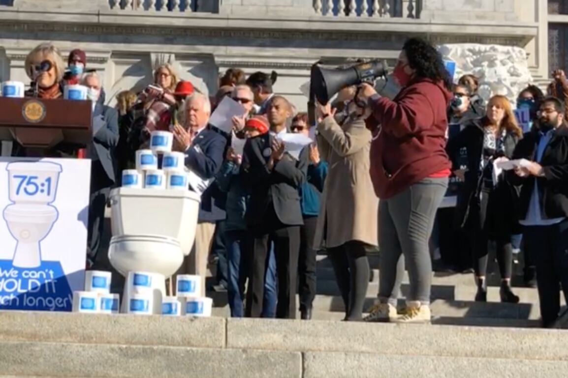 People on the steps of the Pennsylvania Capitol building, with a podium, a toilet and stacks of toilet paper.