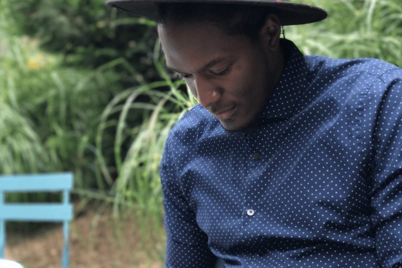 Man in a blue dress shirt and red hat writing in a notebook with a pen.