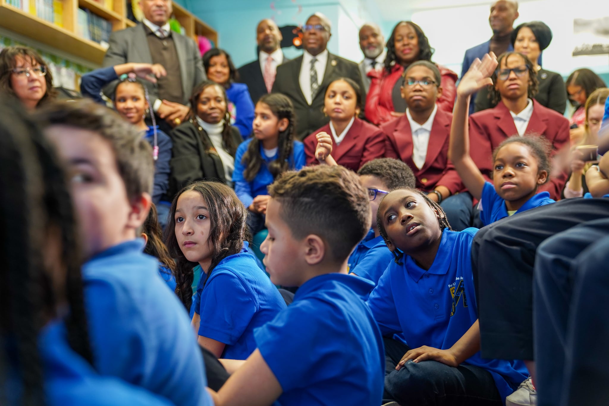 A large group of young students sit in a classroom and listen to someone reading who isn't in the frame.