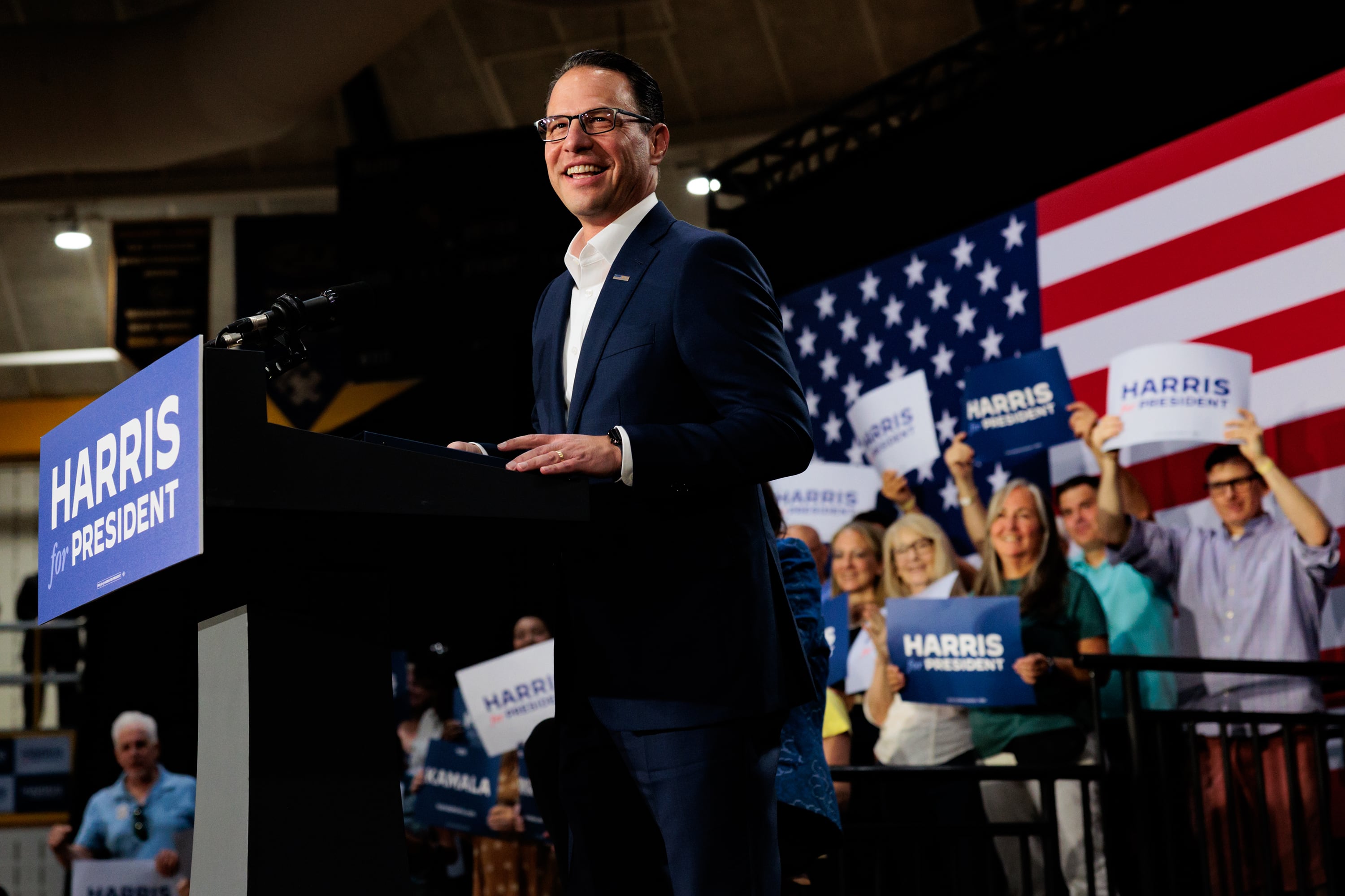 A man wearing a dark blue suit stands behind a podium with a sign that reads "Harris for President" in the front and a group of people standing in the background and in front of a large American flag.