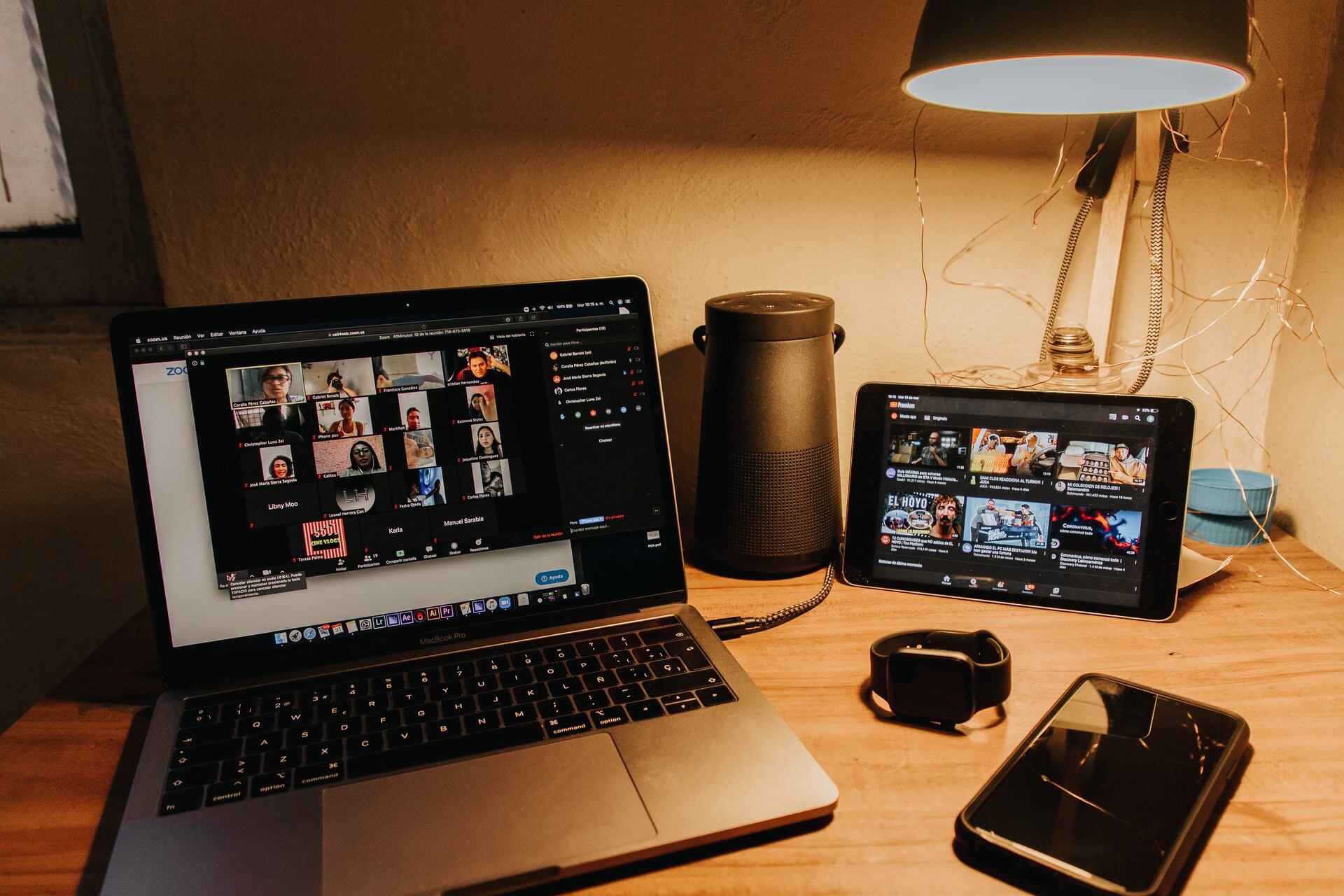 A desk with a laptop, tablet, phone, and watch. On the laptop is an open Zoom conversation with various participants.