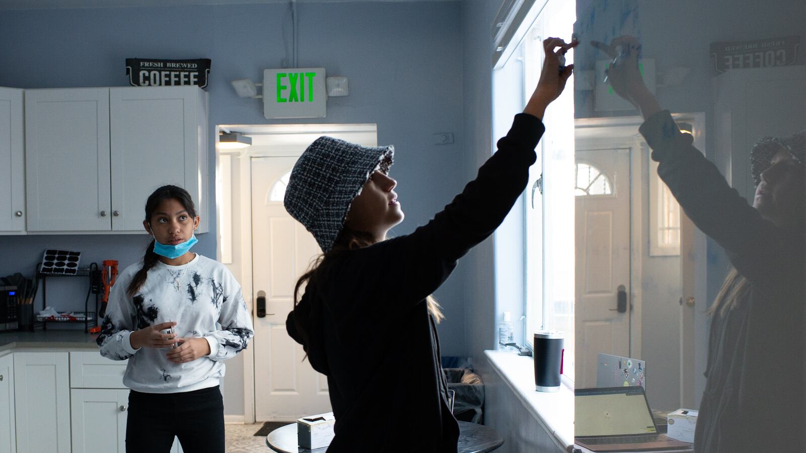 A teenage girl in a bucket hat draws on a white board. Another girl with long dark hair, a mask around her chin, looks on.
