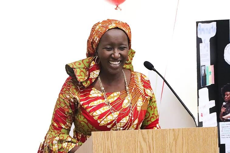 Wearing a colorful dress and scarf, Amnata Sy speaks at a lectern