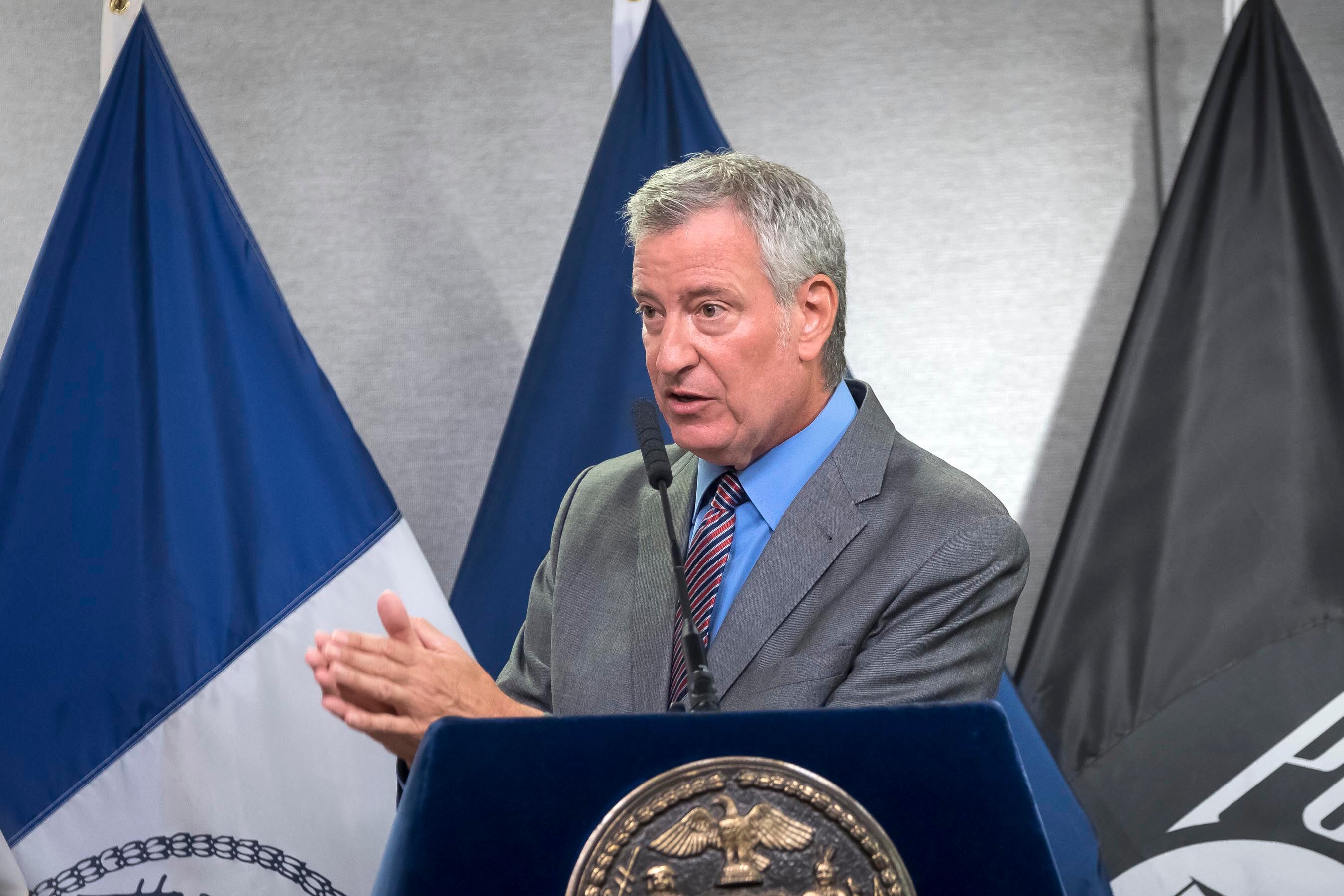 Mayor Bill de Blasio stands at a lectern in front of three flags.