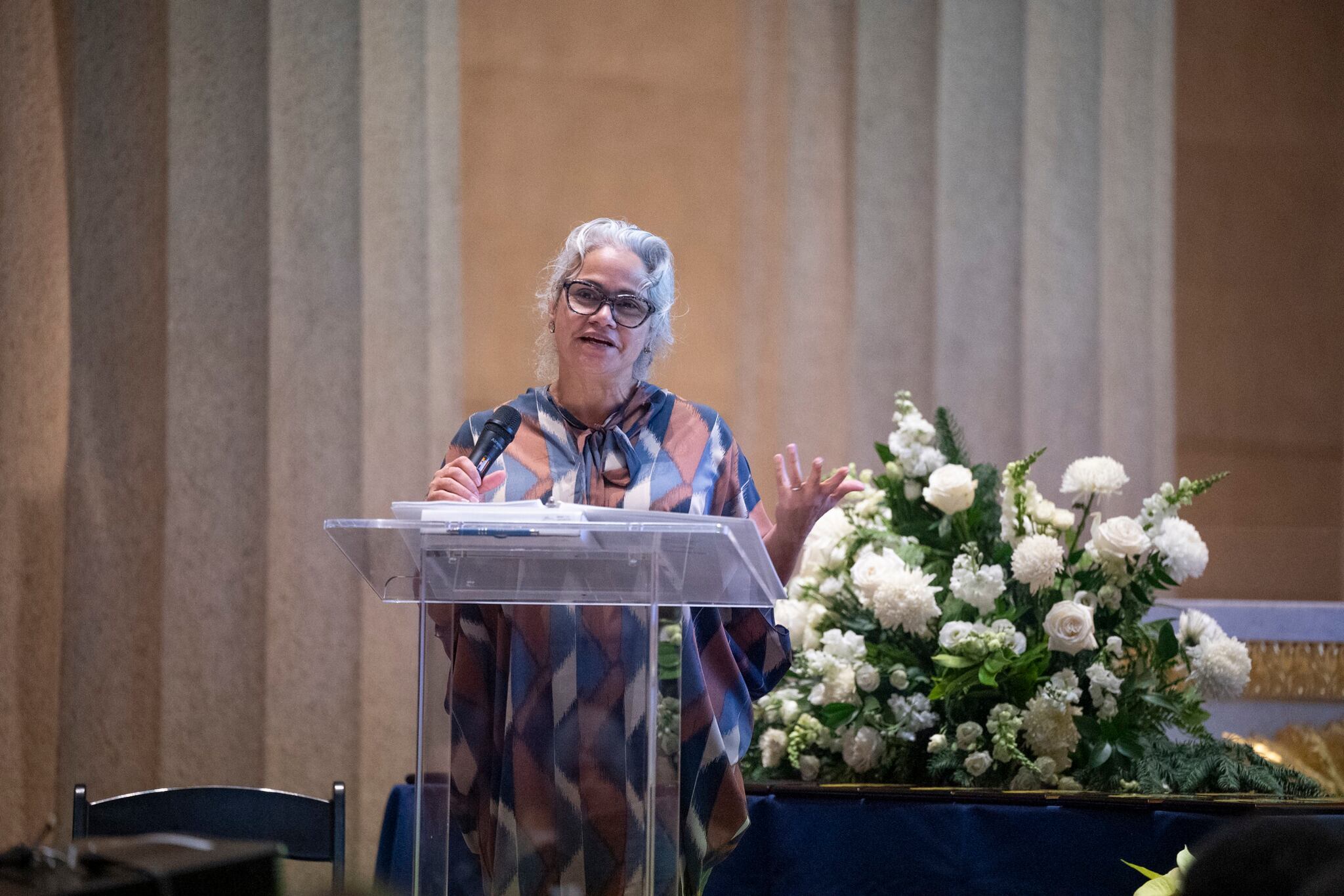 A woman with short white hair and wearing glasses and a colorful blouse stands at a podium with flowers to the right of her and a tan background.