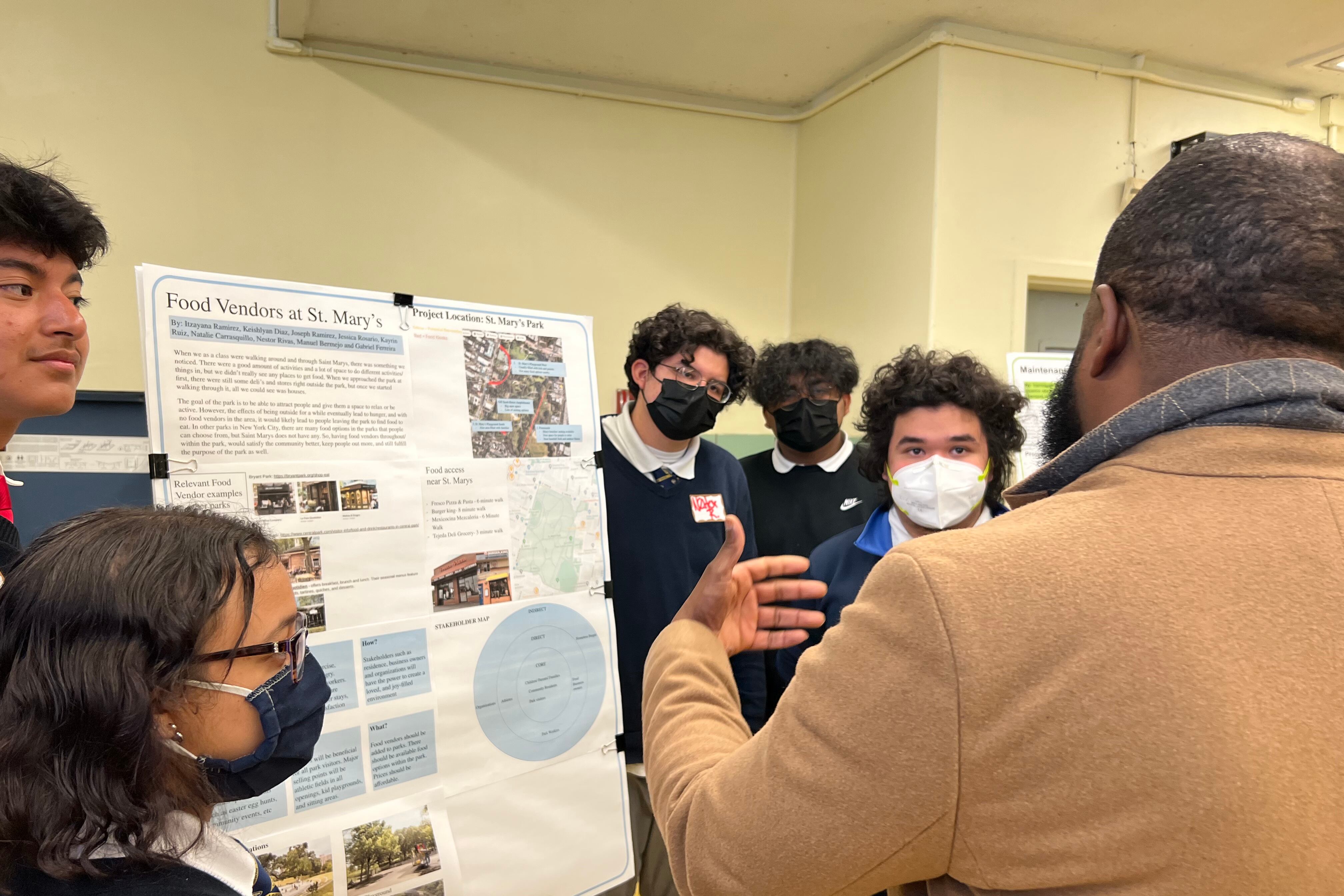 Students gather around a poster board presentation in a room as a man with his back to the camera in a tan coat speaks to them.