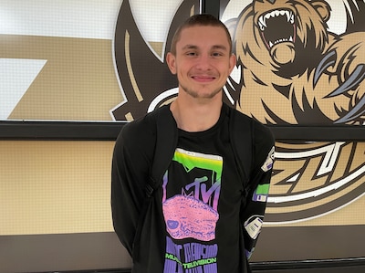 A photograph of a high school senior boy with short hair and wearing a black shirt that has an MTV logo on it and standing against