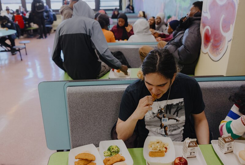 A photograph of an older high school student sitting at a table in a school cafeteria while students sit in the background at various tables.