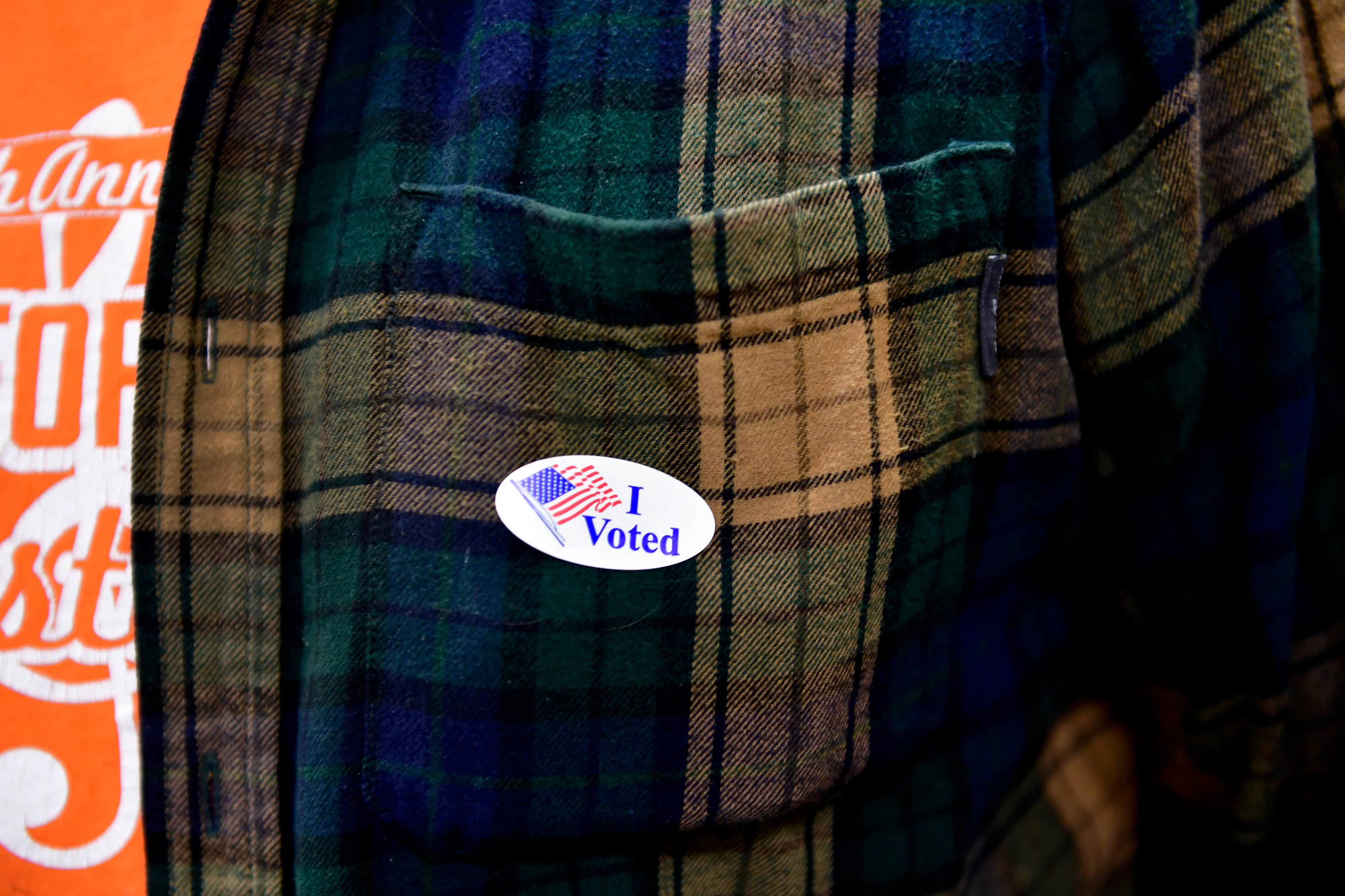 A close-up of an “I Voted” sticker resting on the pocket of a man’s blue, green, and yellow flannel shirt.