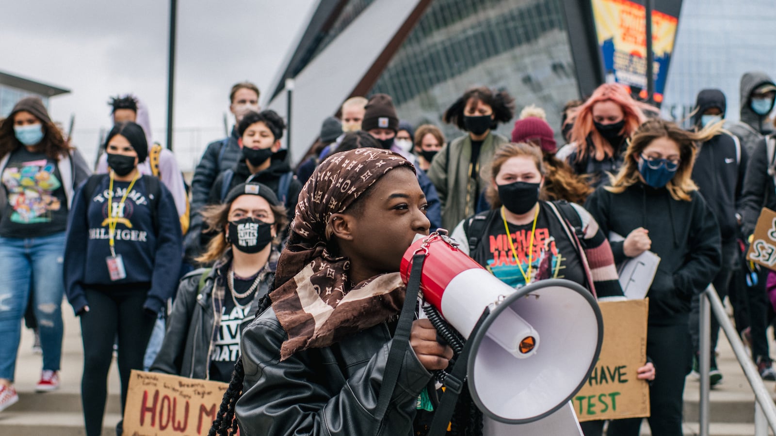 A young woman wearing a black jacket and brown headdress speaks through a red and white bullhorn microphone. There are several students holding signs or standing in solidarity behind her.