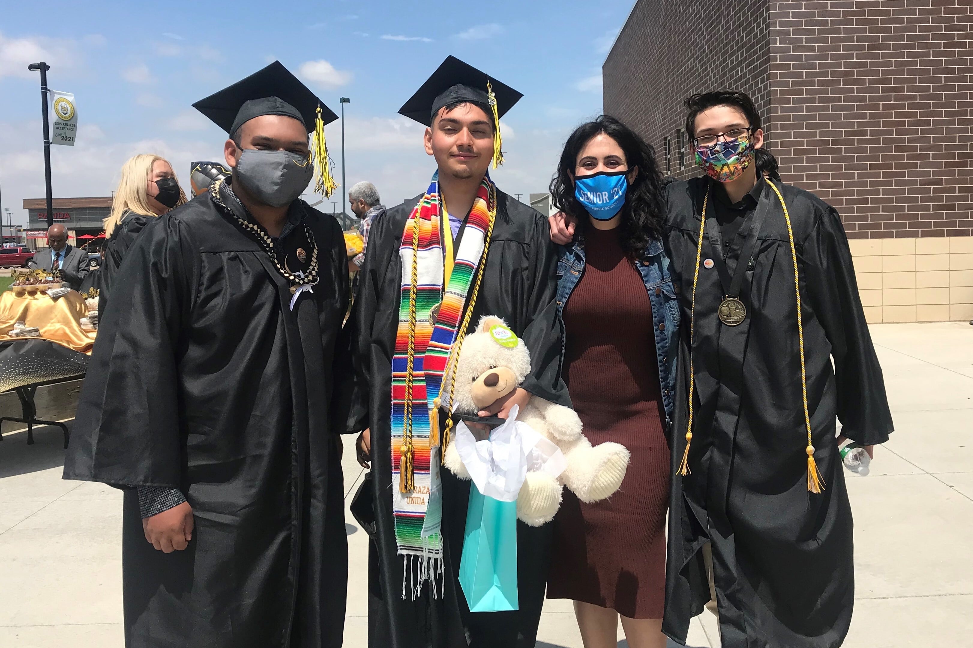 A teacher stands with three high school graduates  outside a building on a sunny day. 