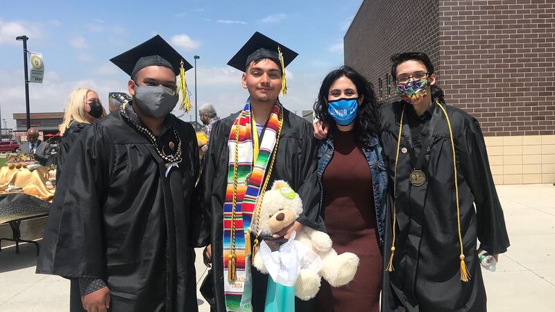 A teacher stands with three high school graduates  outside a building on a sunny day. 