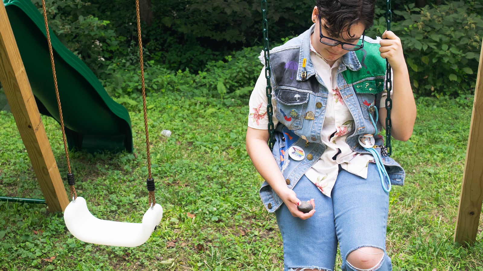 A youth in jeans and a jeans vest sits on a swing and looks downward.