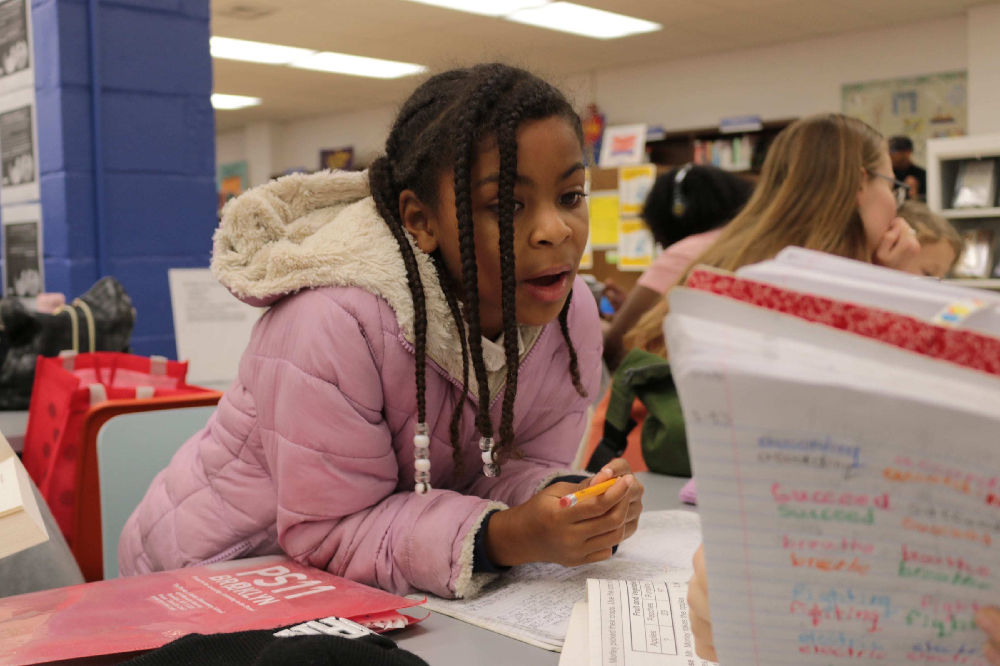 A young girl with long braids and wearing a pink coat reads from a table inside a public library.