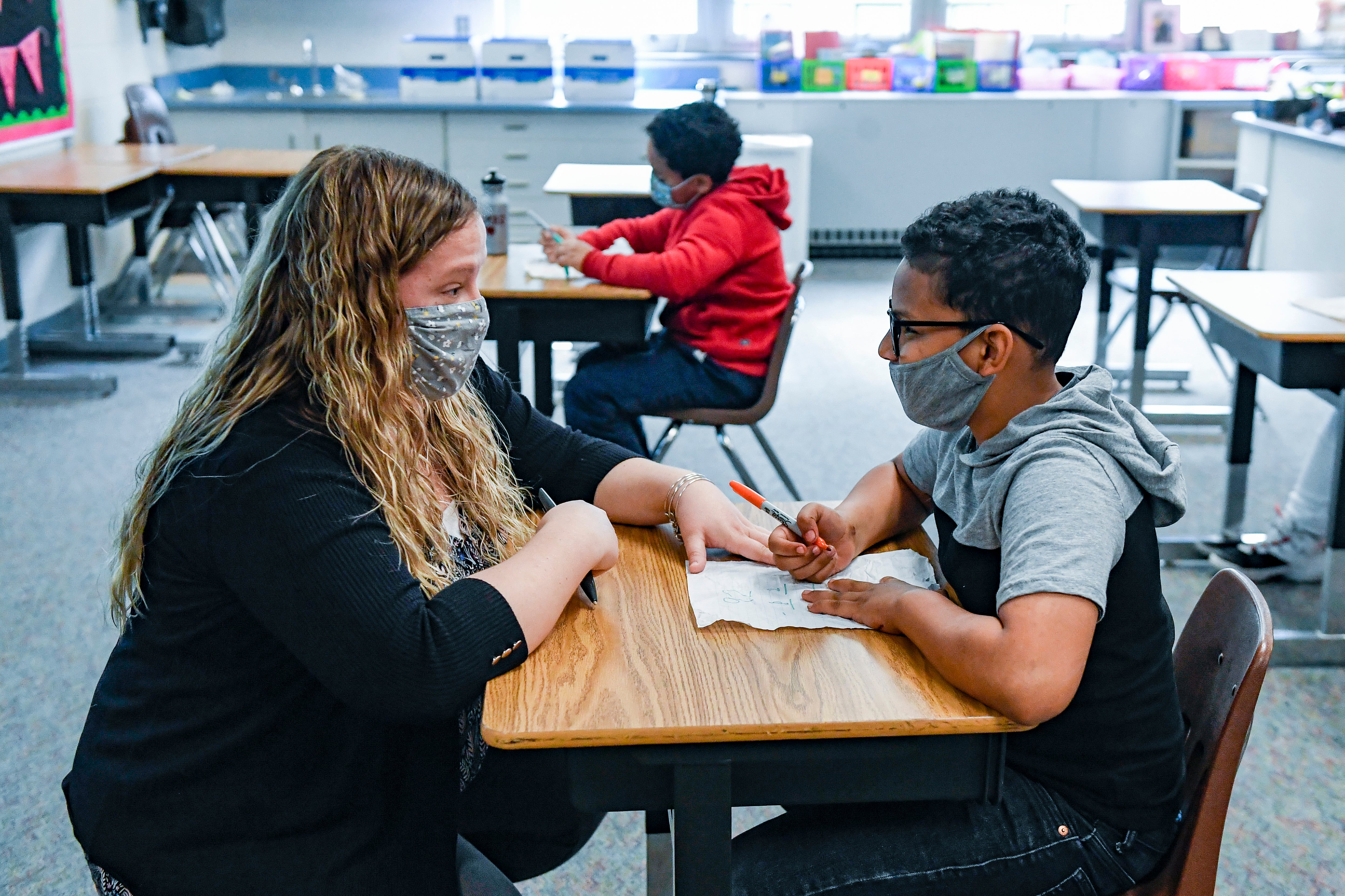 Woman with blond hair wearing a masks sits at a desk across from a male student with glasses holding a pen.