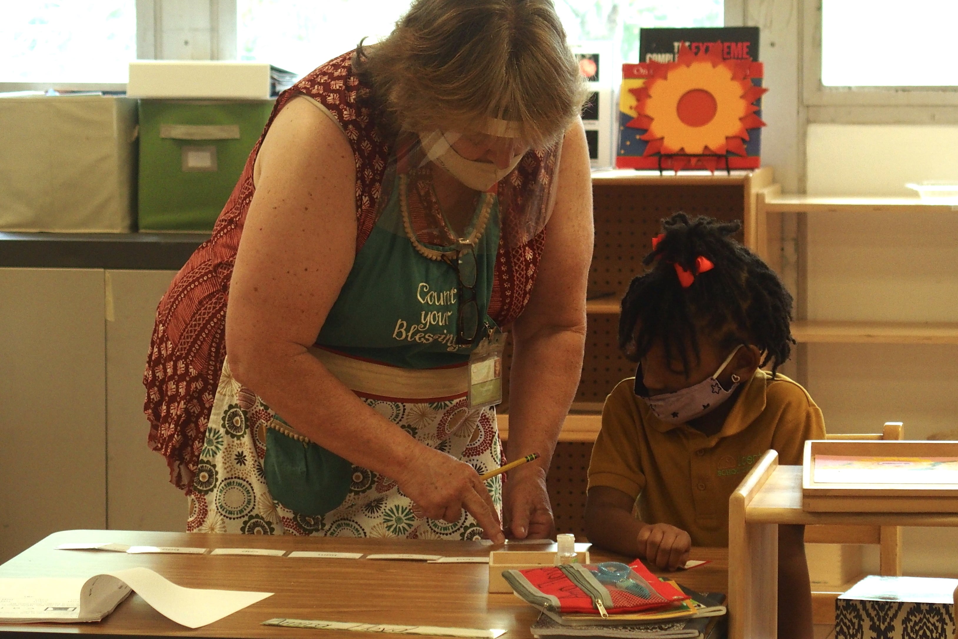 A teacher helps an elementary school student in class while both wear masks