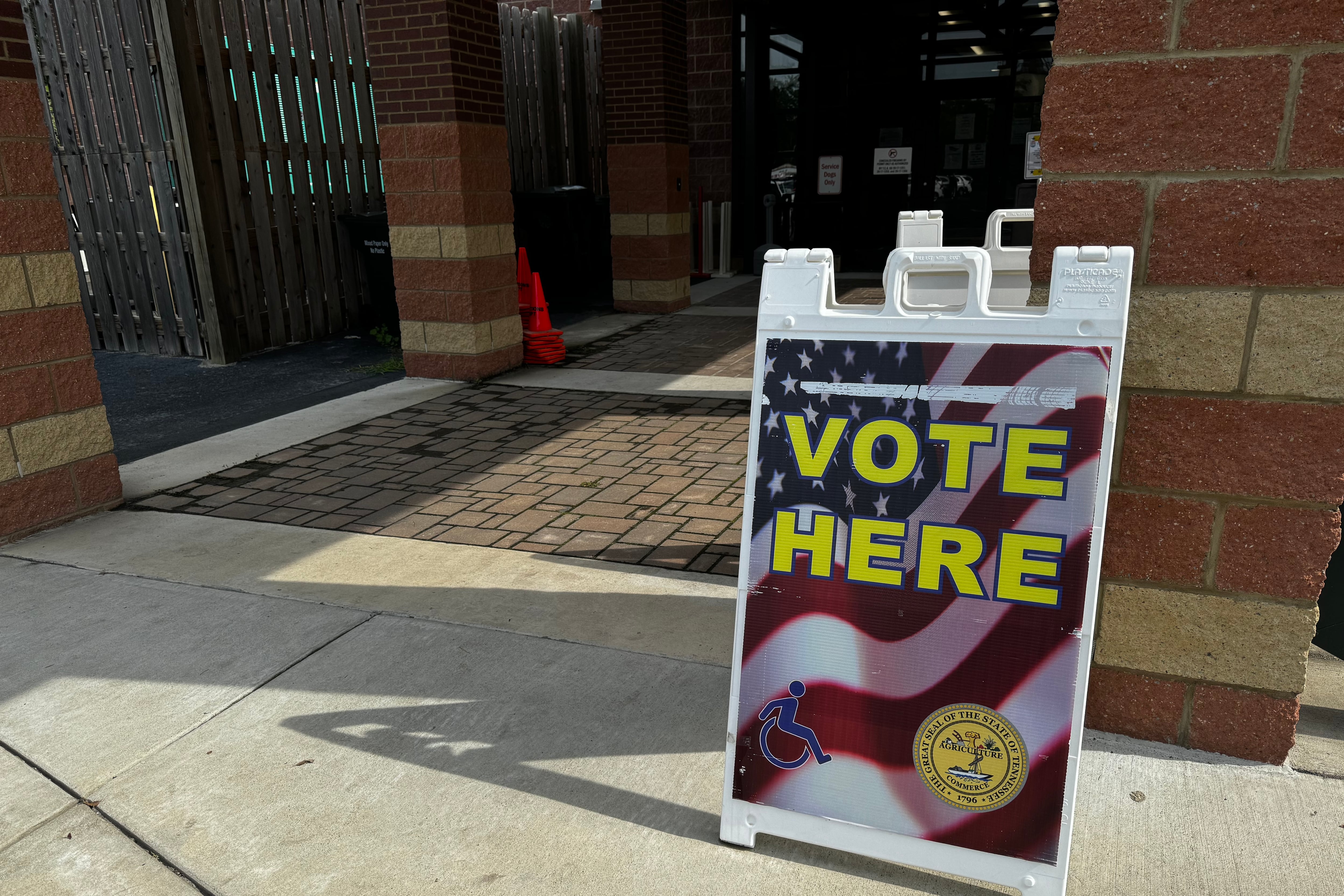 A white plastic sign with an American flag printed on it and the words "vote here" across the middle sits on a concrete sidewalk outside of the entrance to a building.
