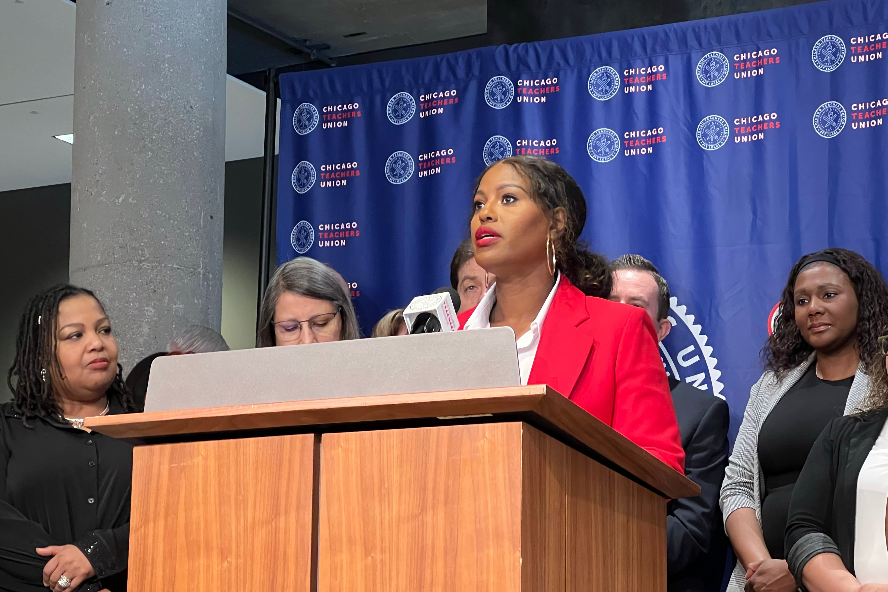 A Black woman wearing a red suit jacket speaks from behind a wooden podium and in front of a blue background an a group of people stand next to her.