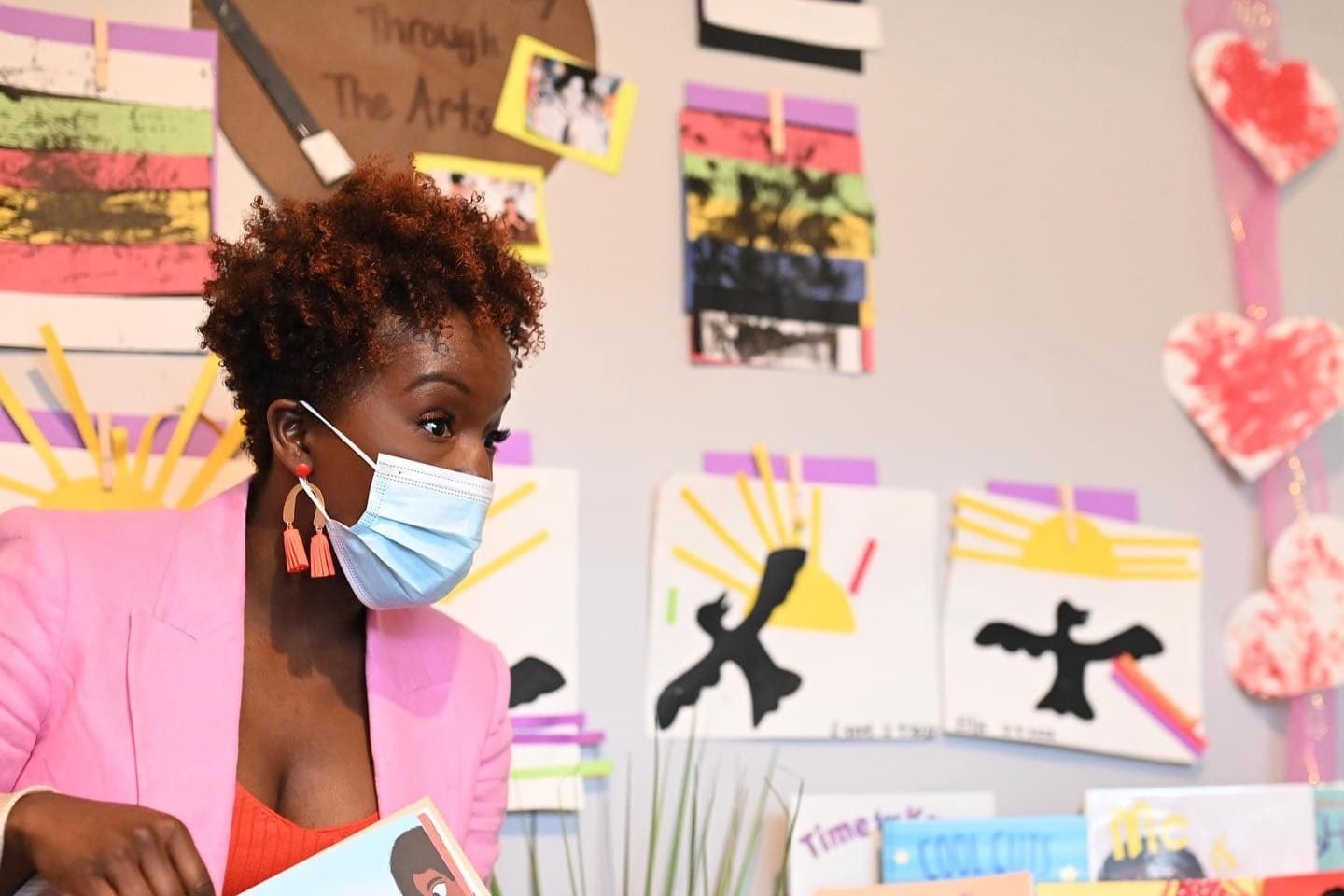 An author in a beautiful pink jacket reads a children’s storybook in a colorful preschool classroom.