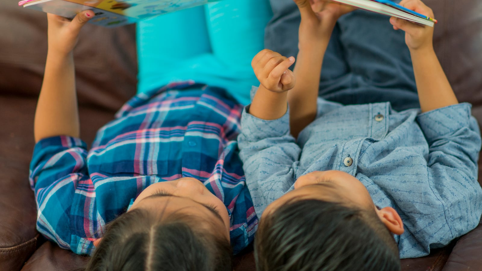 A view from above two children, who are wearing blue shirts and pants, sit on a couch and read a book.