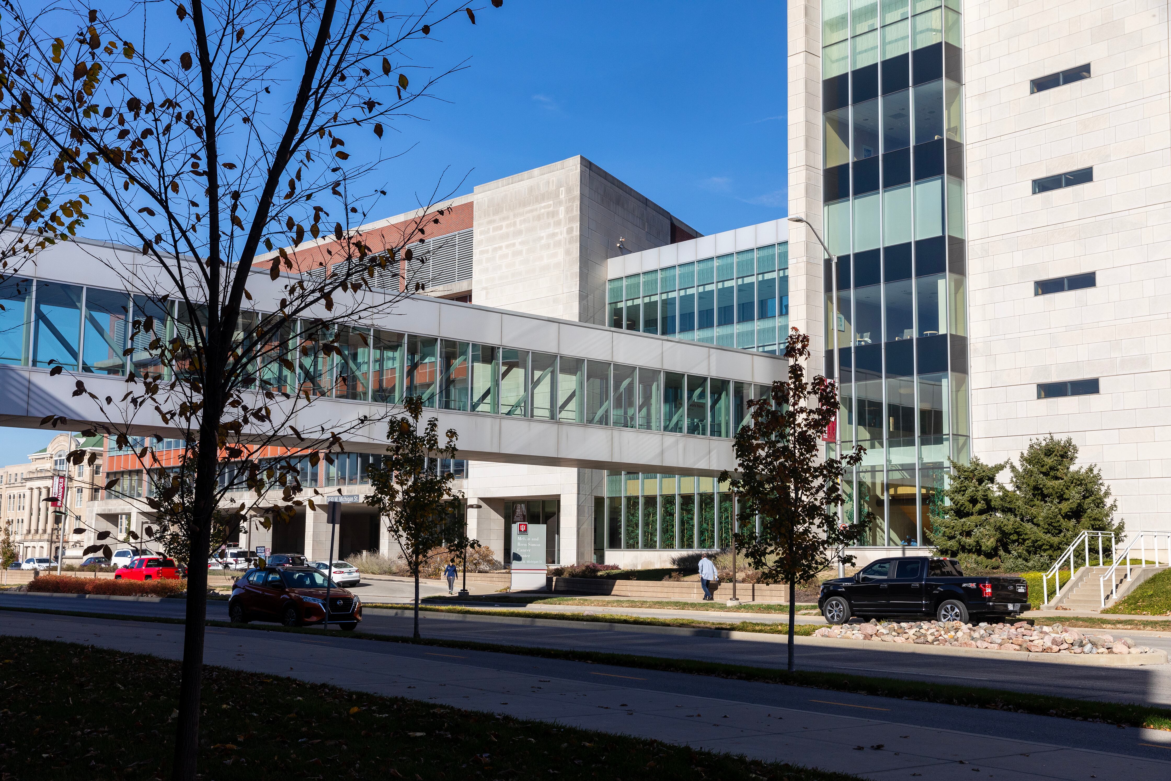 A large white stone building with a glass tunnel connecting to another building. Tree are in the foreground and blue sky in the background.
