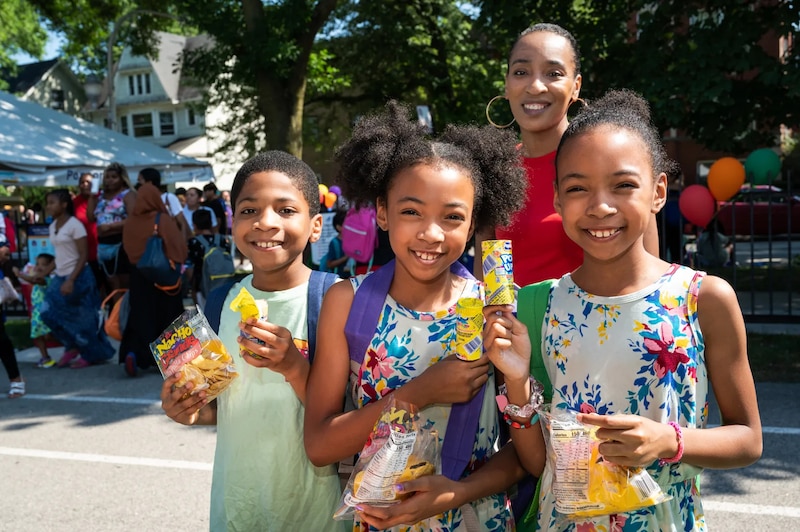 Three young girls smile while wearing new backpacks and enjoying snacks at a back-to-school event.