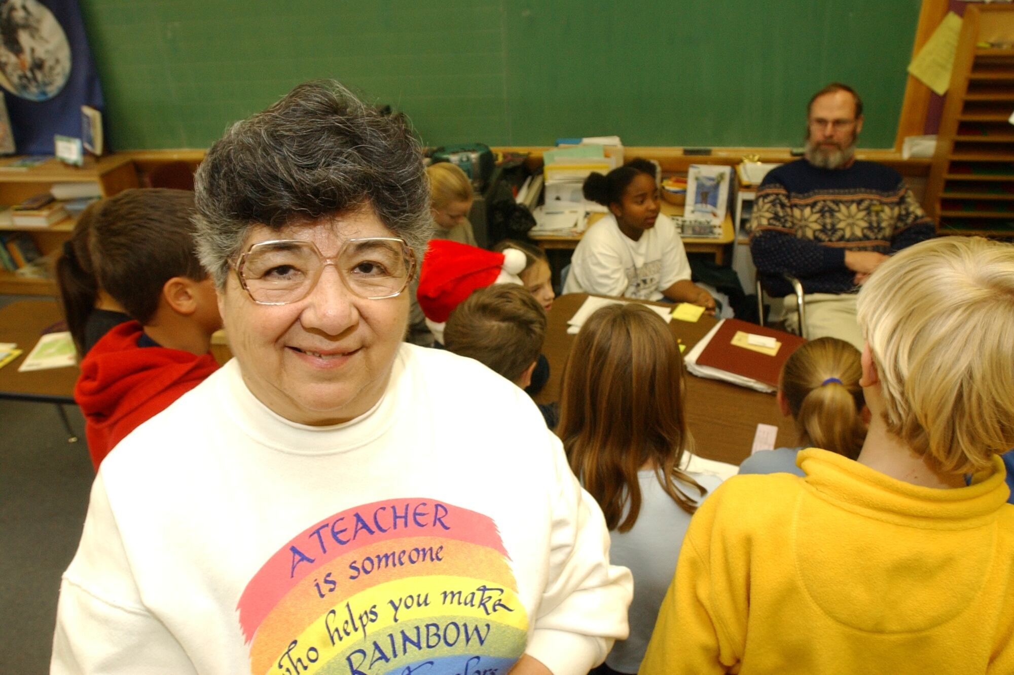 A woman in glasses and a sweatshirt smiles in a classroom. Students can be seen in the background.
