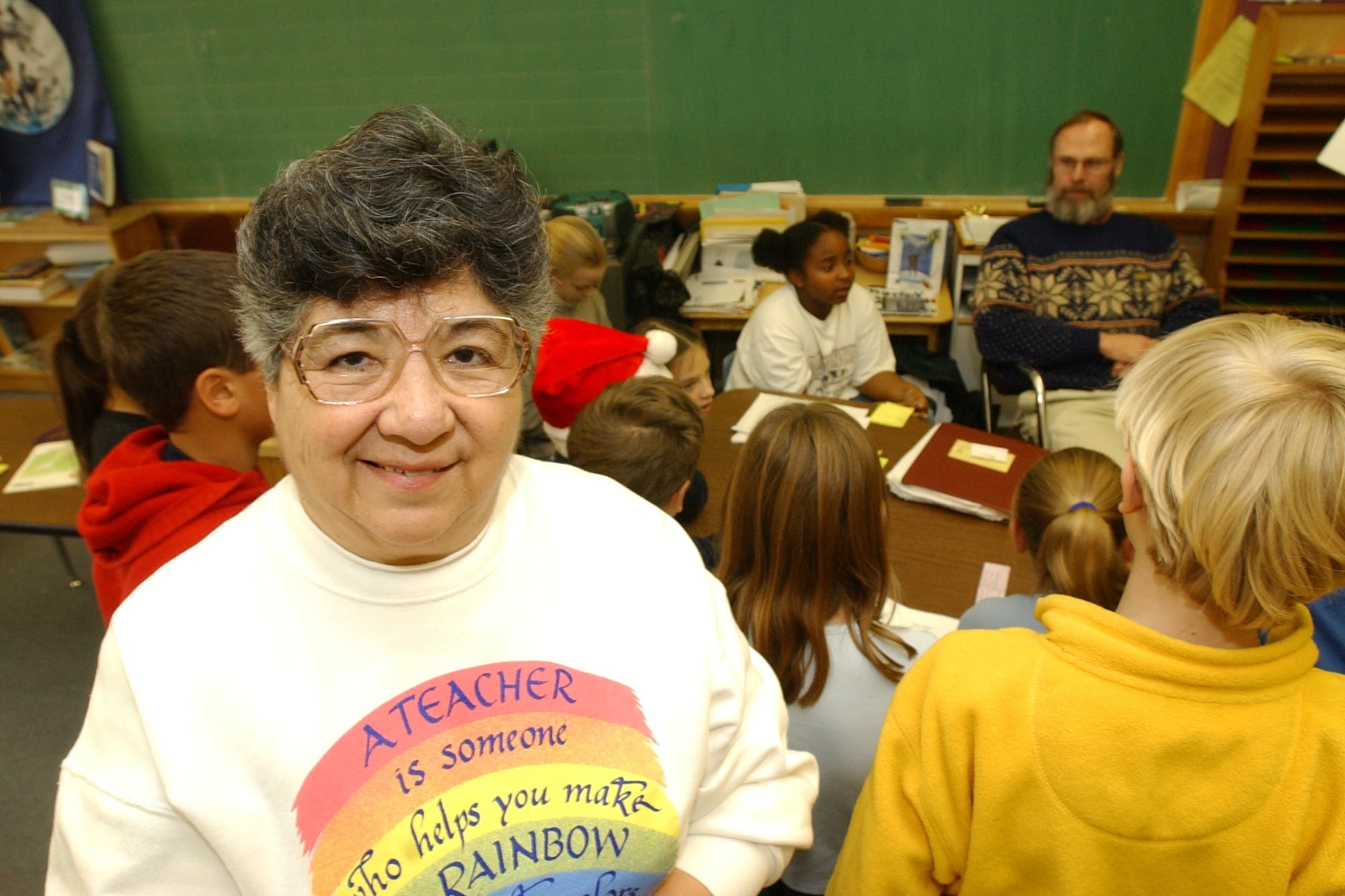 A woman in glasses and a sweatshirt smiles in a classroom. Students can be seen in the background.