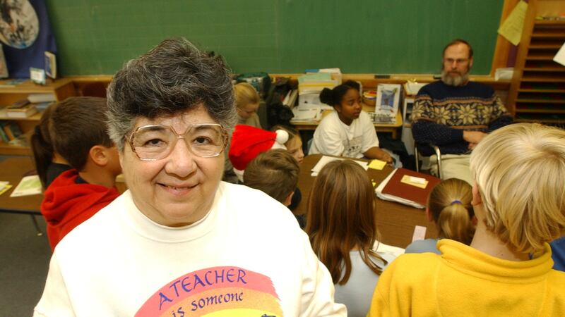 A woman in glasses and a sweatshirt smiles in a classroom. Students can be seen in the background.