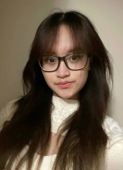 A high school girl with long brown hair and black glasses poses for the camera with a tan background.