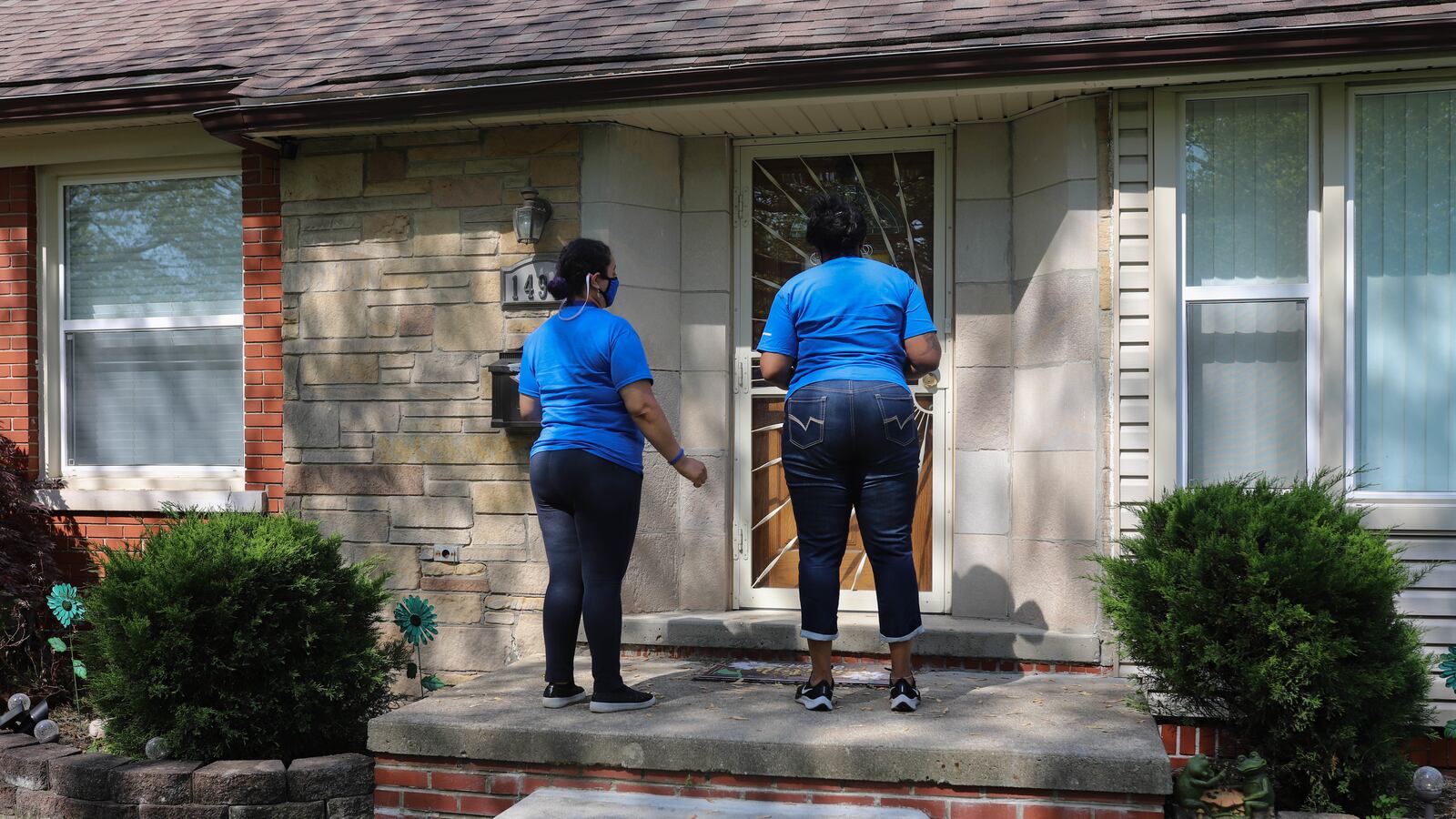 Two women, wearing blue shirts and jeans, stand at the front door of a house in Detroit, Mich. that is patterned from the sun and shadows.