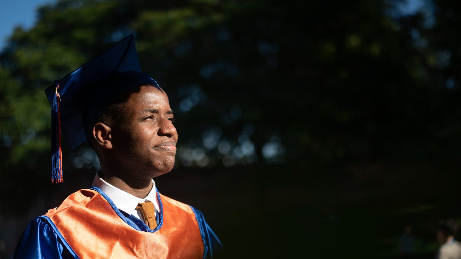 A young man, wearing a blue and orange graduation gown and cap, looks off to the right with a proud smile on his face.