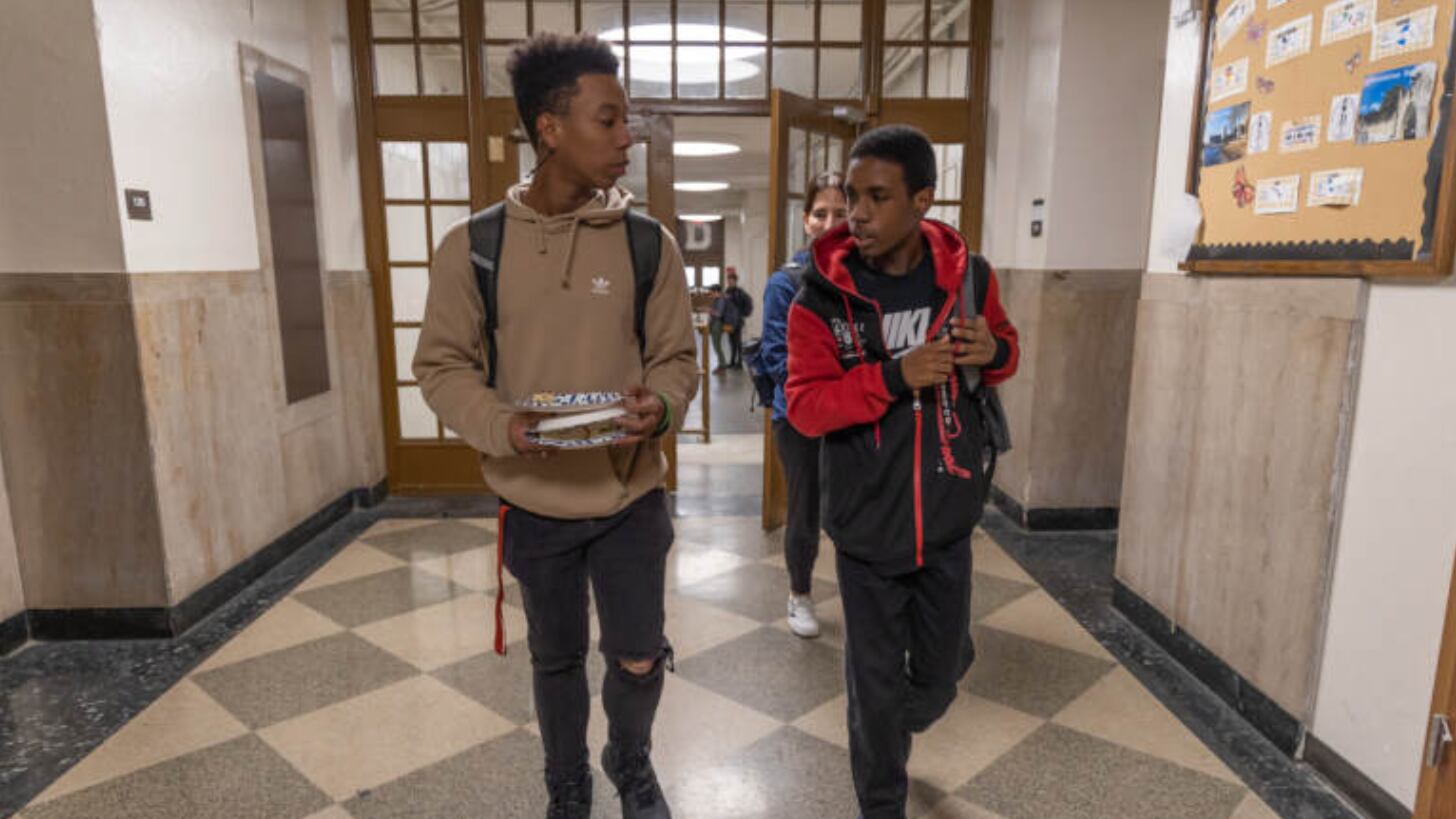 A student wearing a brown hooded sweater and black jeans and black shoes walks next to another student with short hair and a red and black sweater. 